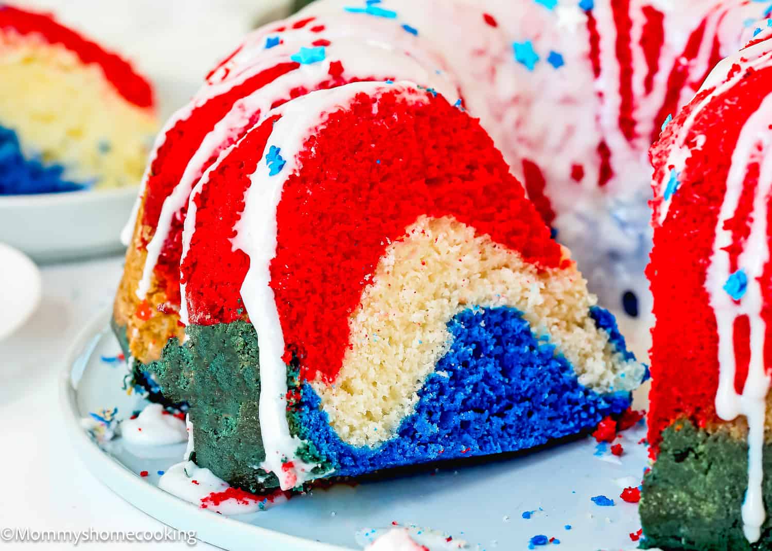 A Red, White & Blue Eggless Bundt Cake for the 4th of July with colorful layers, white icing, and star-shaped sprinkles sits on a plate with a slice cut out.