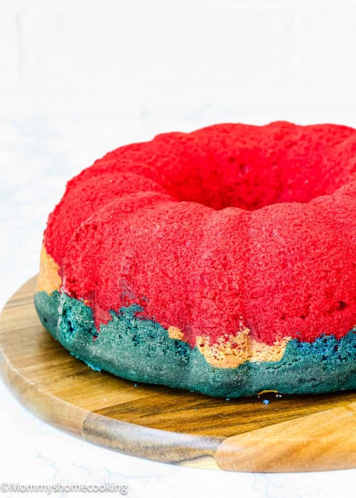 A round bundt cake with a red top, a green base, and yellow accents sits on a wooden board—perfect for celebrations like a White & Blue Eggless Bundt Cake for the 4th of July.