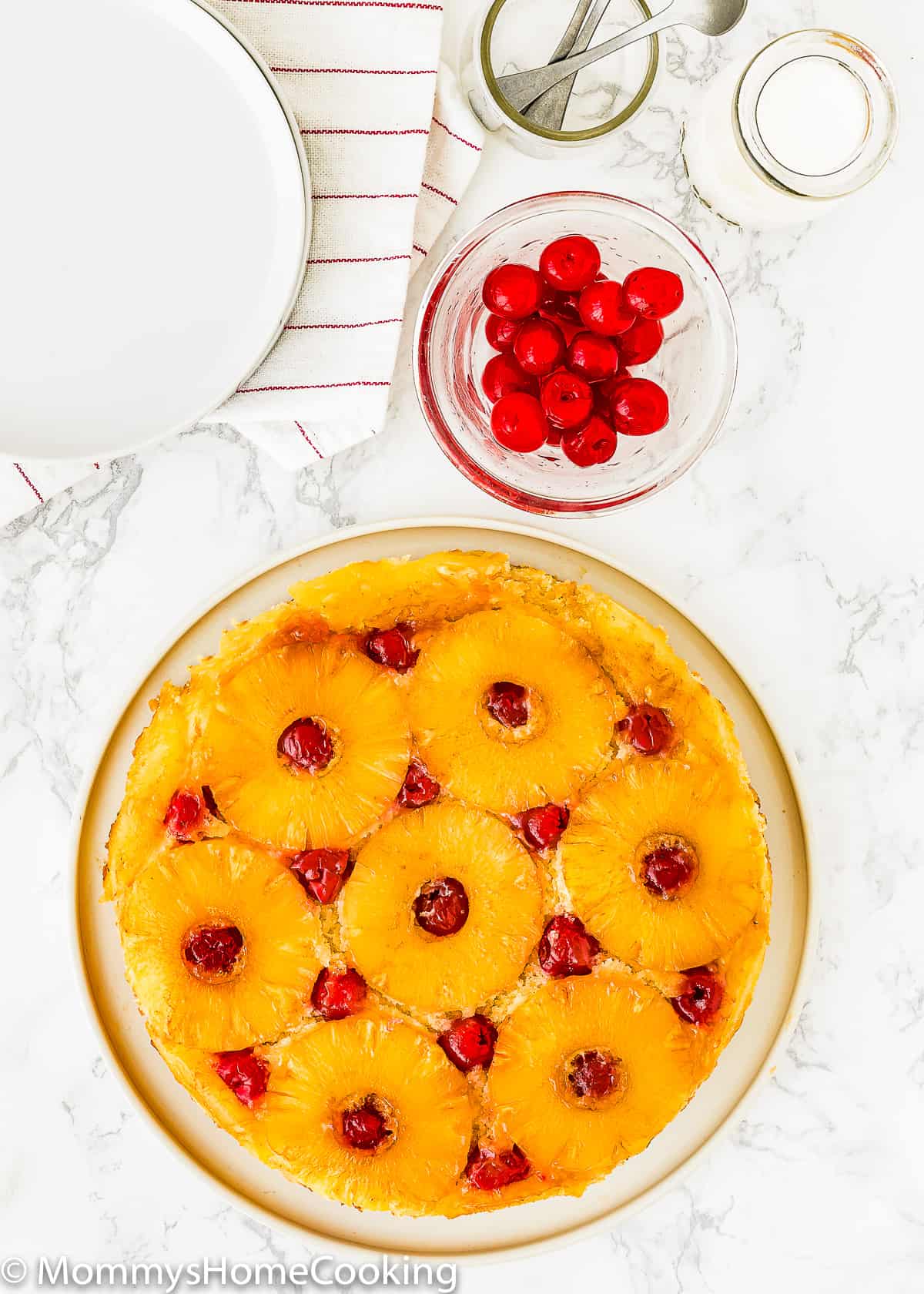 overhead view of an Eggless Pineapple Upside Down Cake on a plate with cherries and plates on the sides.