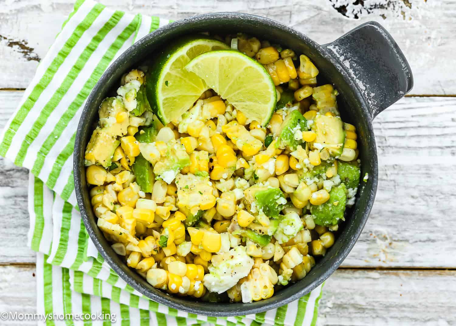 A bowl of Corn Avocado Salad with cheese and lime wedges, placed on a green-and-white striped cloth on a rustic wooden surface.