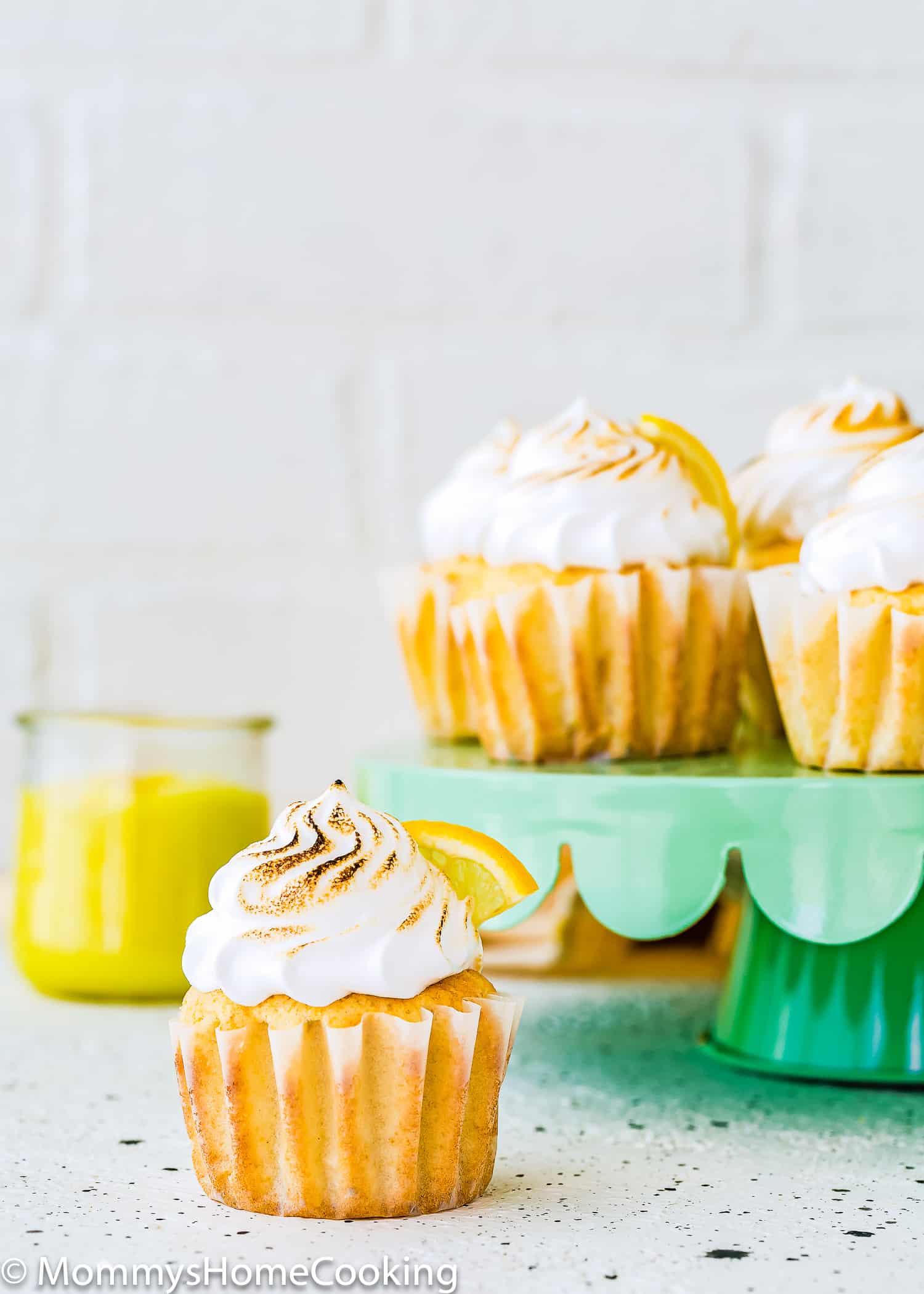 Eggless Lemon Meringue Cupcake on a green cake stand with a container in the background with egg-free lemon curl.