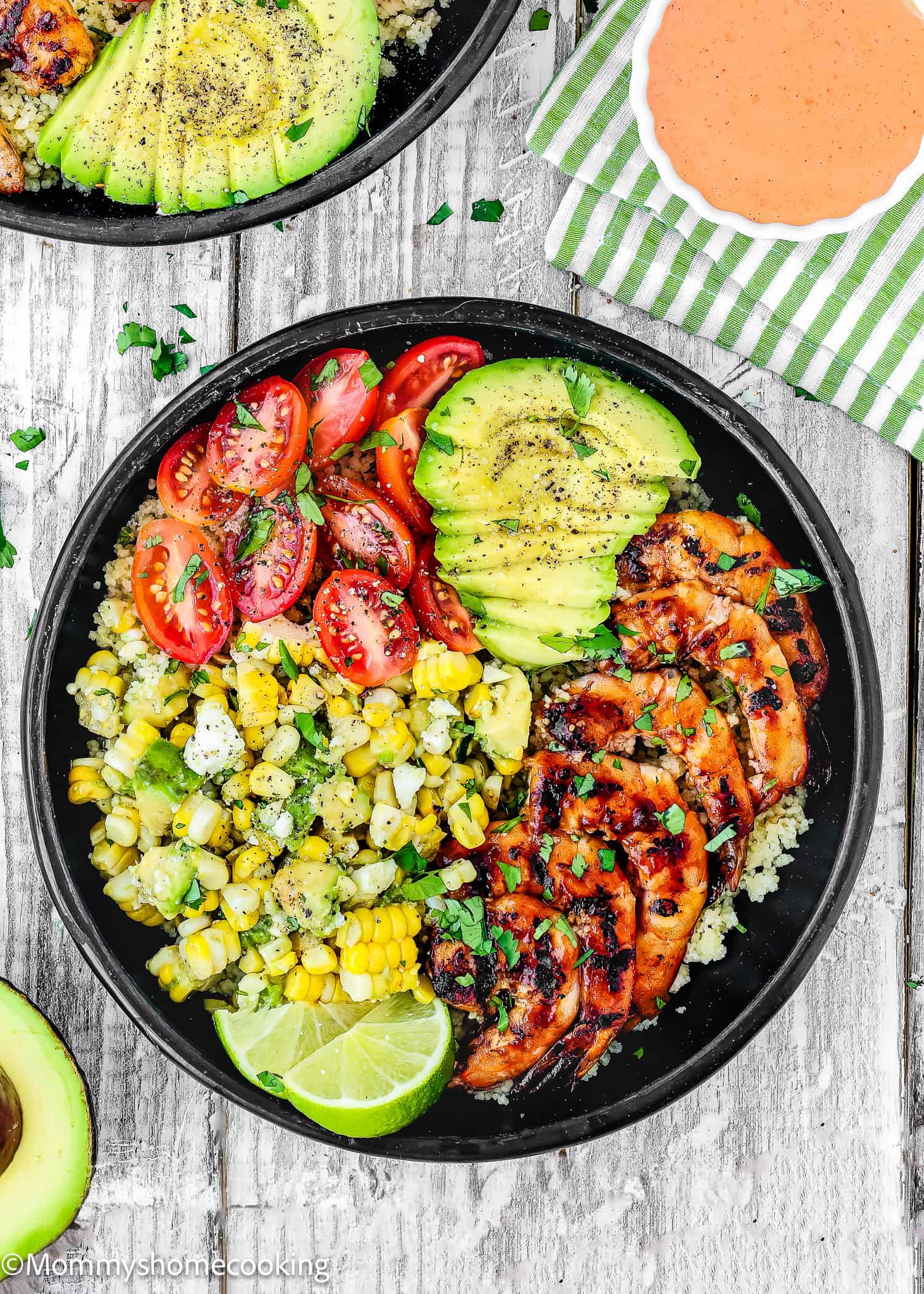 A black bowl filled with grilled shrimp, sliced avocado, cherry tomatoes, corn, lime, and herbs on a bed of grains, sits on a rustic white wooden surface.