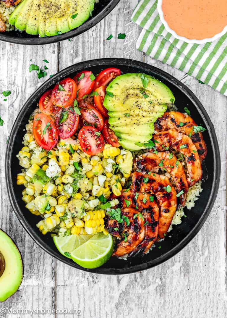 A black bowl filled with grilled shrimp sliced avocado, cherry tomatoes, corn, lime, and herbs on a bed of grains, sits on a rustic white wooden surface.