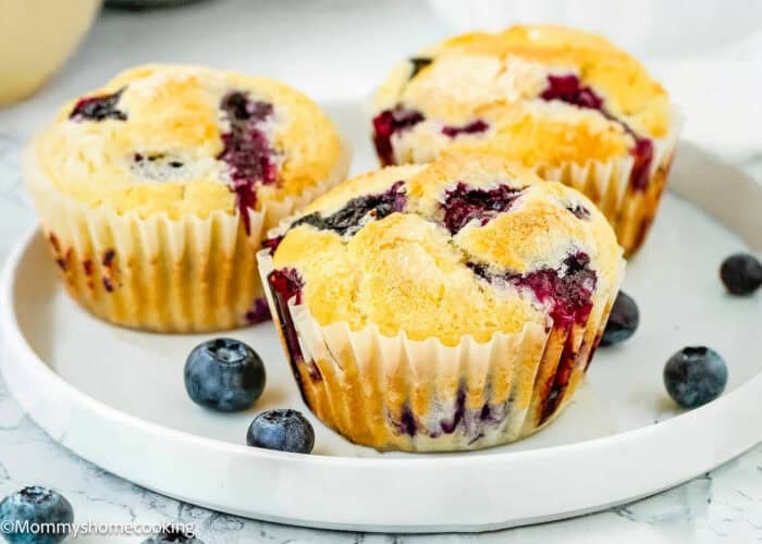 Three Eggless Blueberry Muffins sit on a white plate with a few fresh blueberries scattered around them.
