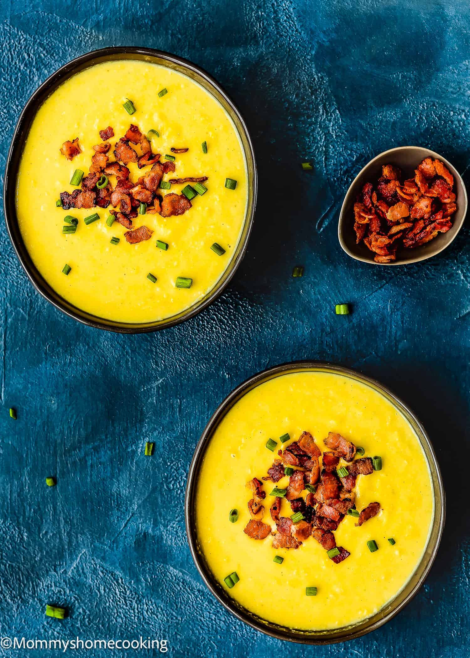 Two bowls of Easy Creamy Corn Soup, topped with chopped bacon and chives, are served on a dark blue surface, with an extra bowl of bacon on the side.