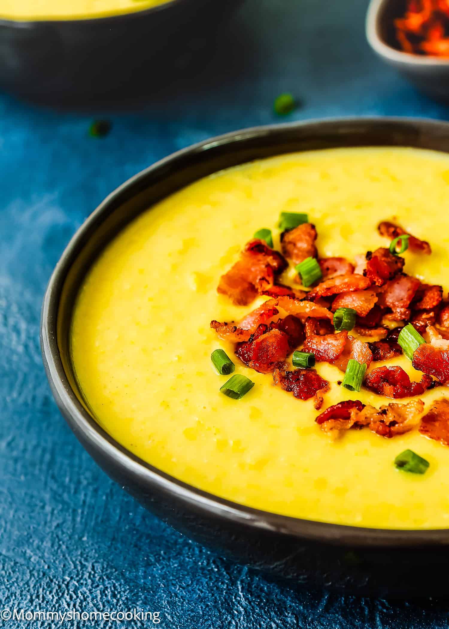 A bowl of Easy Creamy Corn Soup topped with crispy bacon pieces and chopped green onions, set on a blue surface.