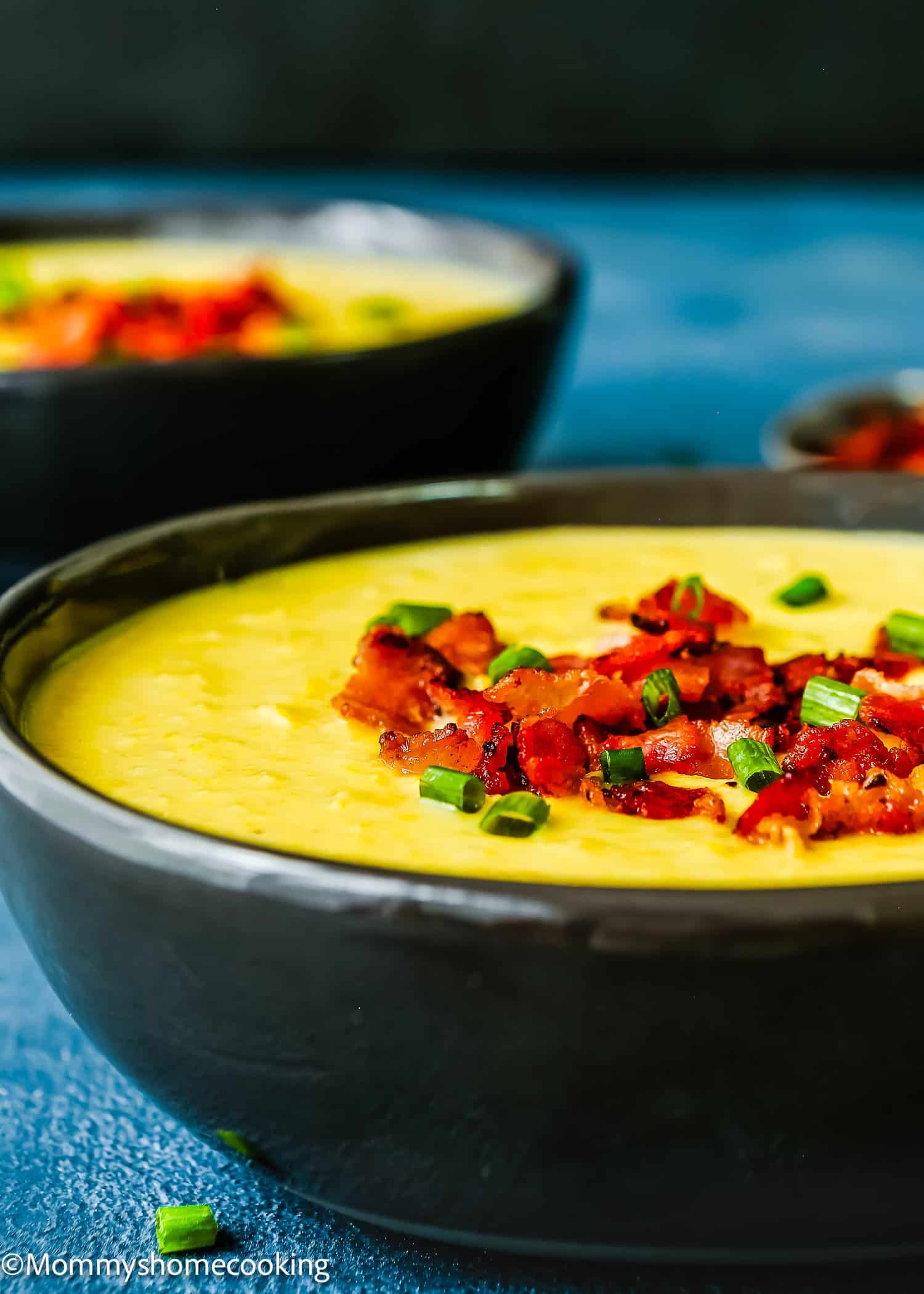 A bowl of Easy Creamy Corn Soup, topped with crispy bacon pieces and chopped green onions, with another bowl and a small dish in the background.