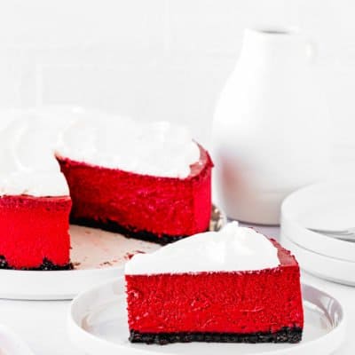 a sliced of Eggless Red Velvet Cheesecake on a plate with the whole cheescake in the background,