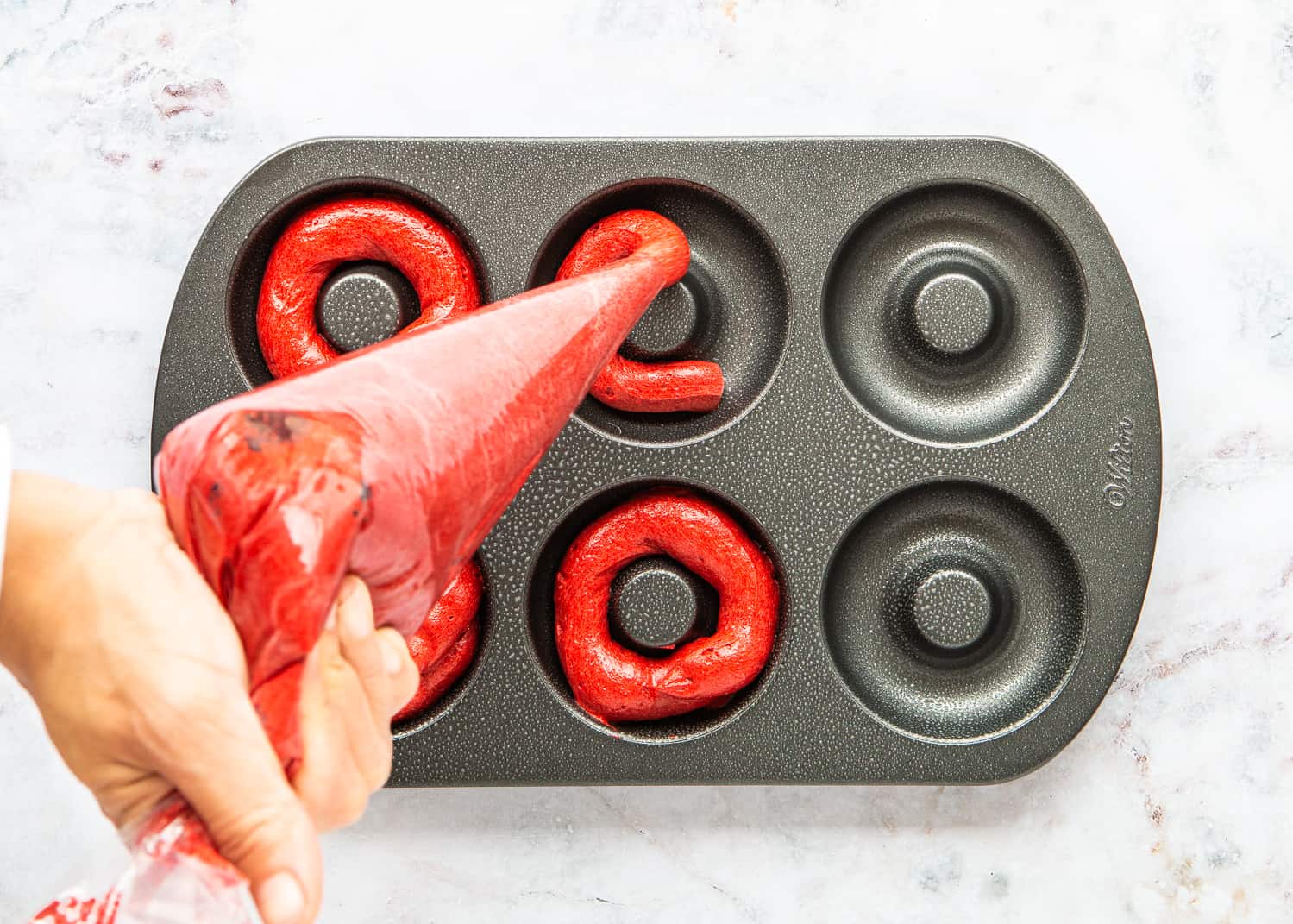 a hand holding a piping bag filled with Eggless Red Velvet Donuts batter and filling a donut pan.