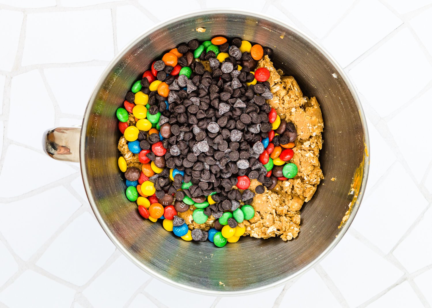 Eggless Monster Cookies dough in a stand mixer bowl with chocolate chips and m&ms on top. 