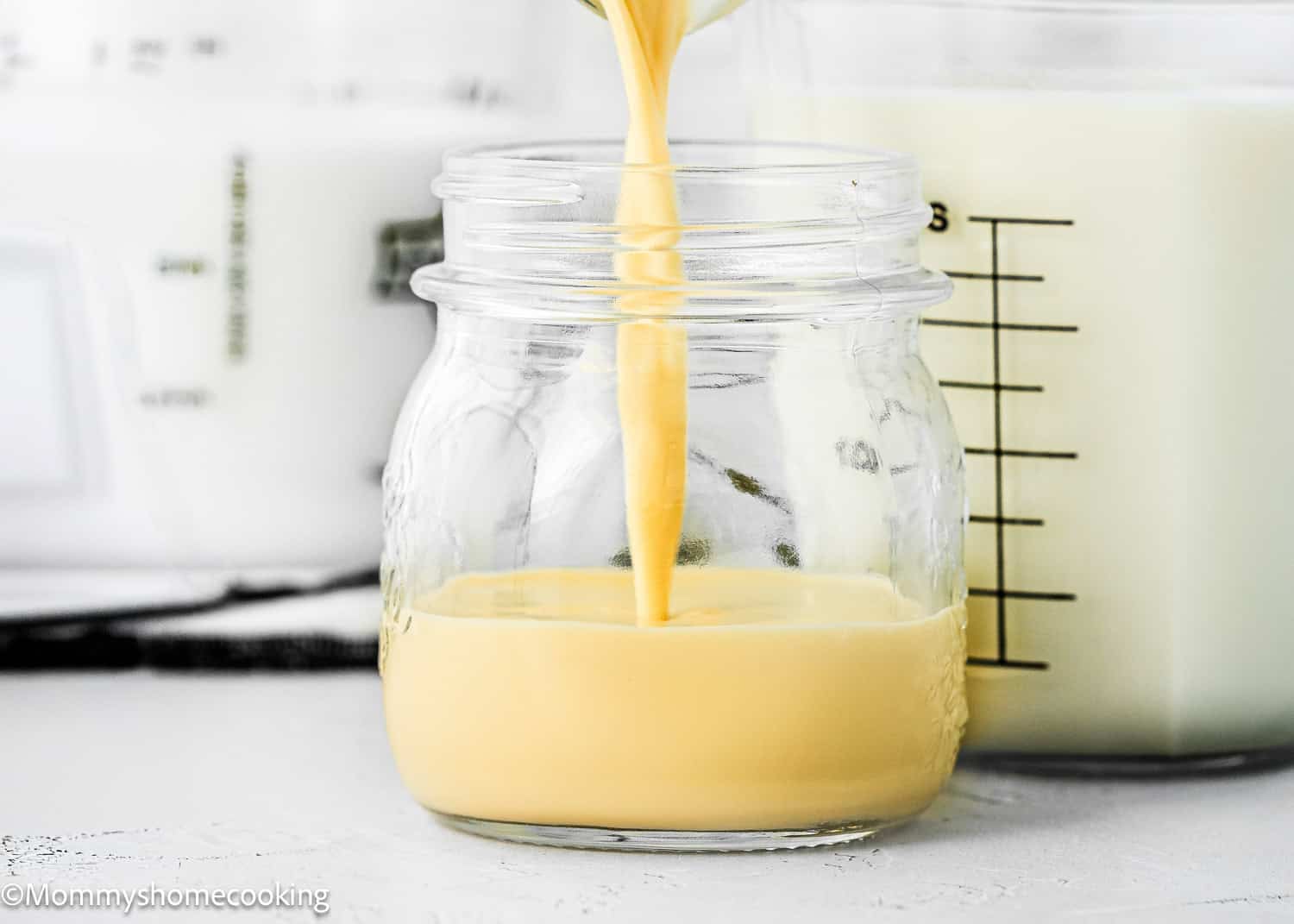 A thick yellow liquid is being poured into a small glass jar, hinting at the process of how to make evaporated milk, with a container of milk visible in the background.