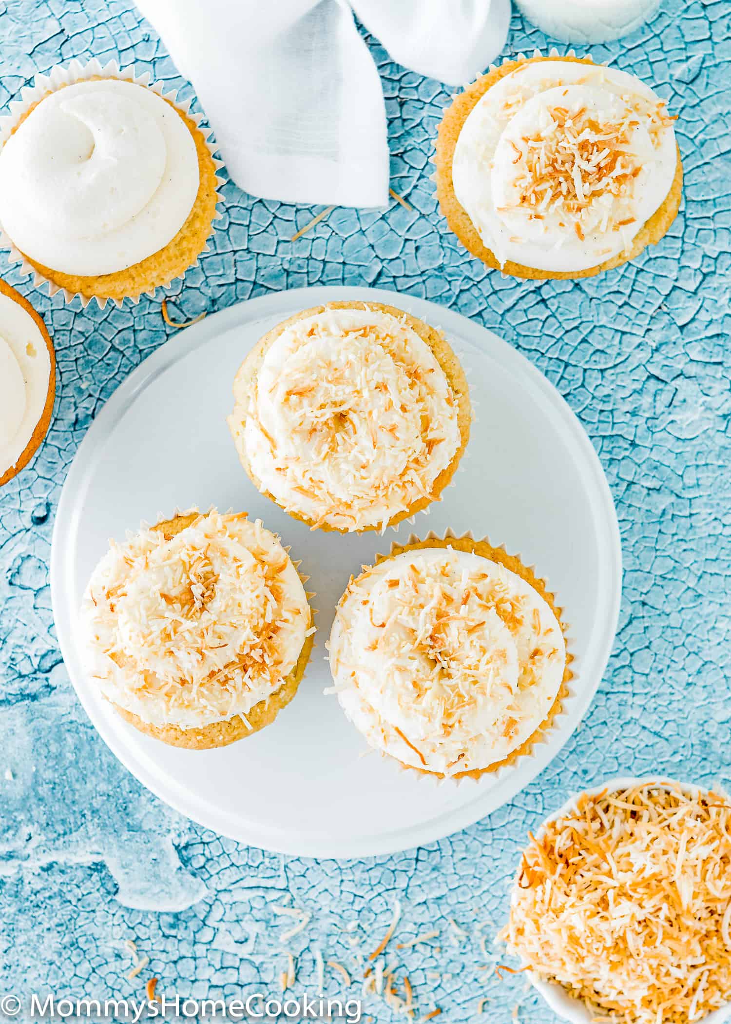overview of a plate with three eggless coconut cupcakes with frosting and coconut on top. 