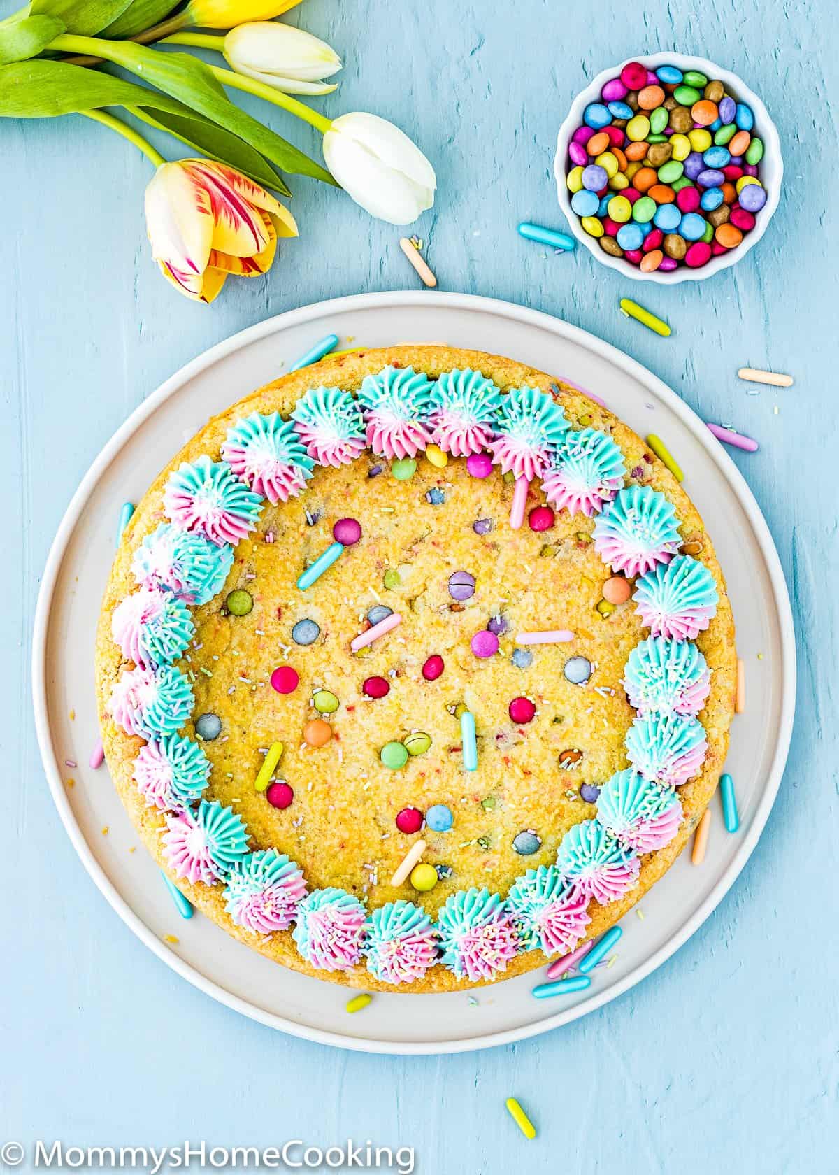 Eggless Easter Sugar Cookie Cake decorated with buttercream and sprinkles on a blue surface with flowers and chocolate candies in a bowl