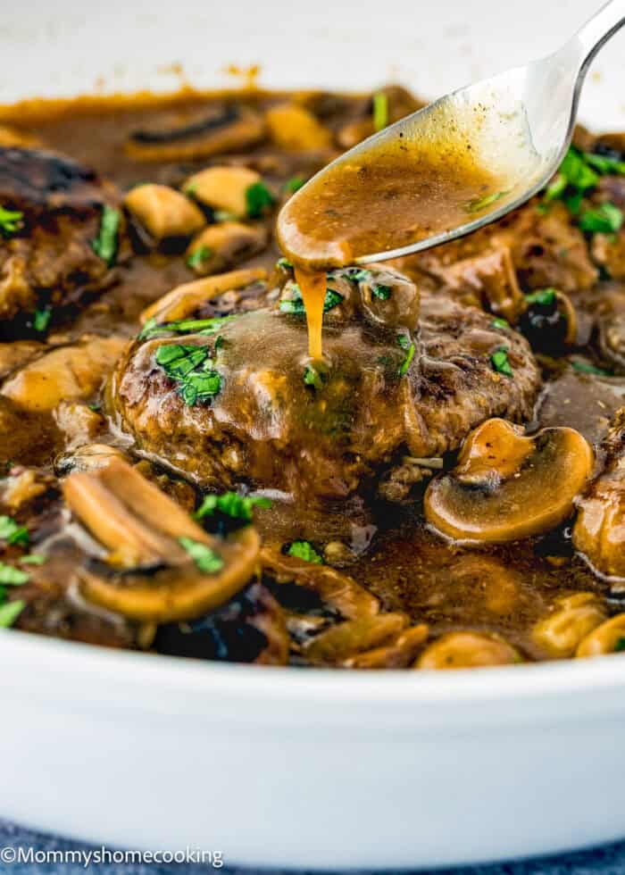 A spoon drizzles brown gravy over an eggless Salisbury steak with mushrooms and fresh herbs in a white skillet.