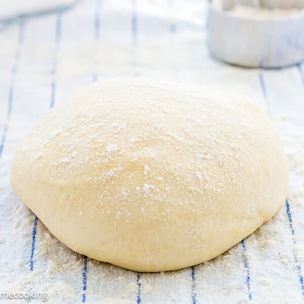 Ball of raw dough dusted with flour resting on a striped cloth, perfect for your favorite homemade pizza dough recipe, with a round metal cutter in the background.