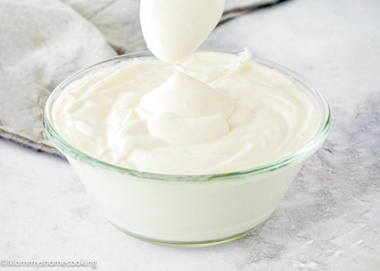 A glass bowl filled with thick, creamy white yogurt sits ready, while a spoon above holds a dollop—perfect for learning how to make sour cream at home.
