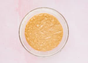 A glass bowl filled with light tan Eggless Strawberry Banana Bread batter sits on a pale pink surface.