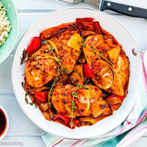 A skillet with The Juiciest Peri Peri Chicken Breasts (One Skillet), cooked with bell peppers, lemon slices, and herbs, sits next to a bowl of white rice and a small bowl of sauce on a white wooden surface.