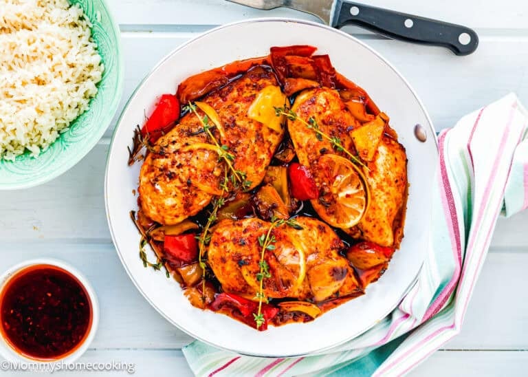 A skillet with The Juiciest Peri Peri Chicken Breasts (One Skillet), cooked with bell peppers, lemon slices, and herbs, sits next to a bowl of white rice and a small bowl of sauce on a white wooden surface.