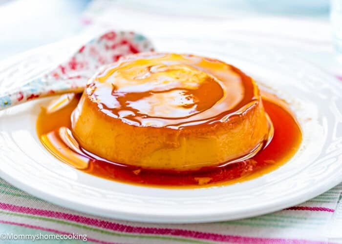 A Venezuelan Flan Quesillo dessert sits on a white plate with caramel sauce and a floral-patterned spoon beside it.