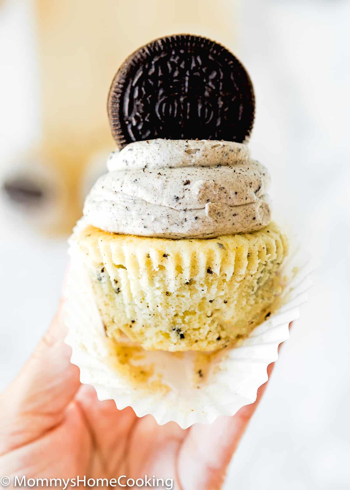 a hand holding an eggless oreo cupcake, frosted with oreo buttercream, and topped with a oreo cookies. 