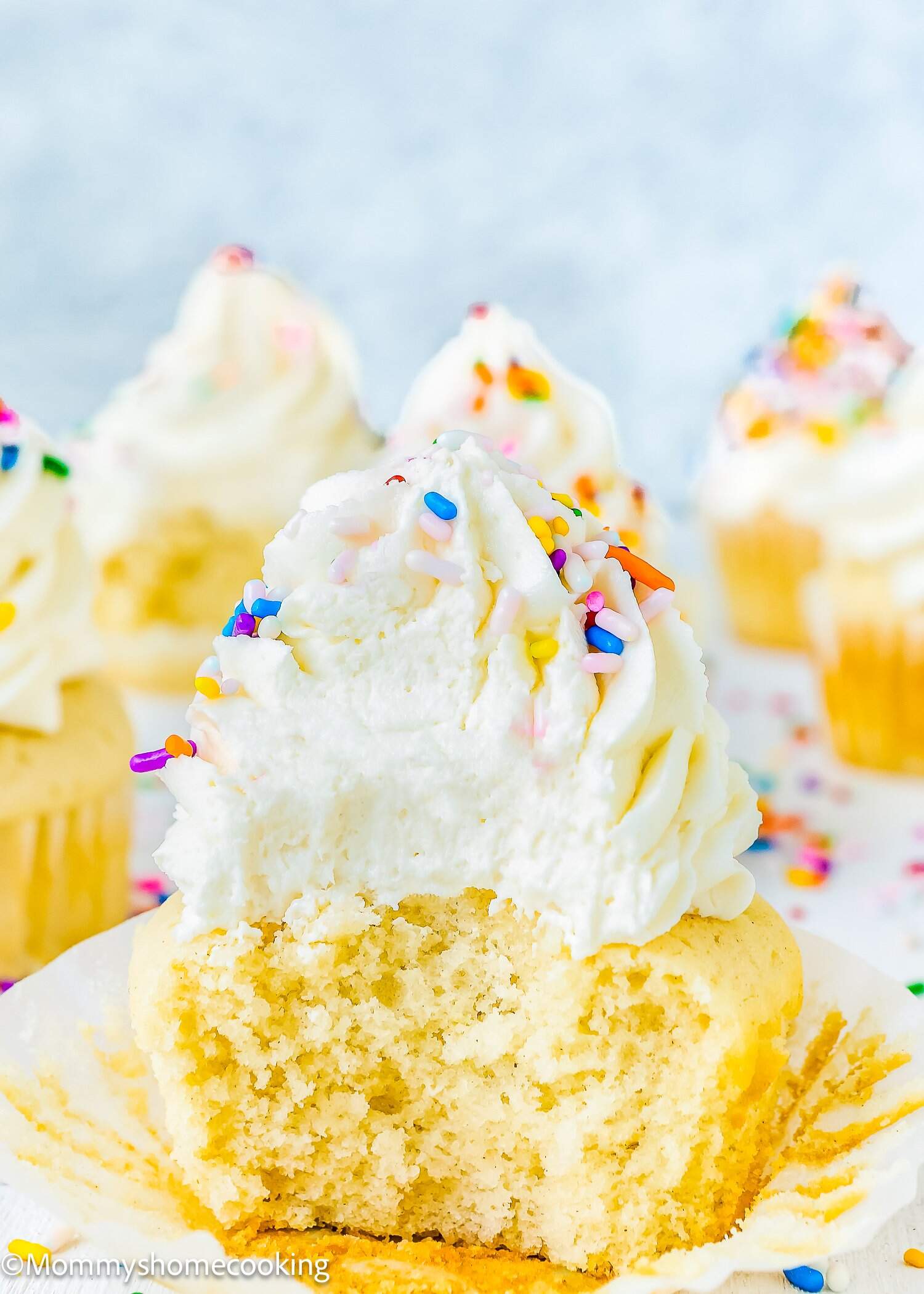 An eggless vanilla cupcake with white frosting and colorful sprinkles, partially eaten, sits on a white surface with more eggless vanilla cupcakes in the background.