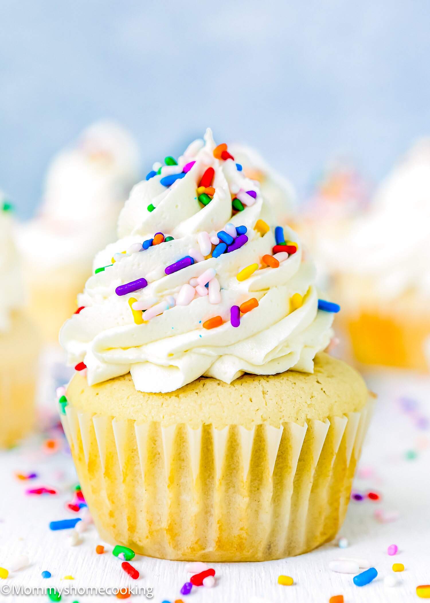 Eggless Vanilla Cupcakes with swirled white frosting and colorful sprinkles, surrounded by similar cupcakes and scattered sprinkles on a white surface.