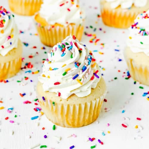 Eggless Vanilla Cupcakes topped with swirls of white frosting and colorful sprinkles are arranged on a white surface with scattered sprinkles.