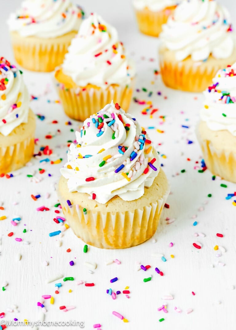 Eggless vanilla cupcakes topped with swirls of white frosting and colorful sprinkles are placed on a white surface and sprinkled with sprinkles.