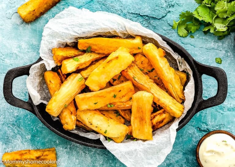 A black dish filled with crispy fried yuca sticks on parchment paper, garnished with chopped herbs, shows exactly How to Make Yuca Fries, complete with a small bowl of dipping sauce and fresh cilantro on the side.