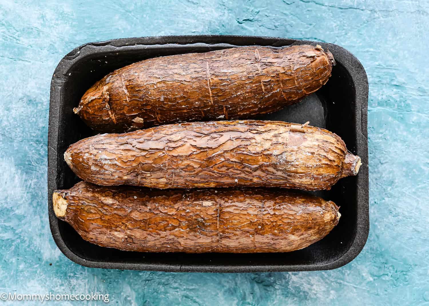 Three whole baked cassava roots, perfect for learning how to make yuca fries, are placed side by side in a black rectangular baking dish on a blue textured surface.