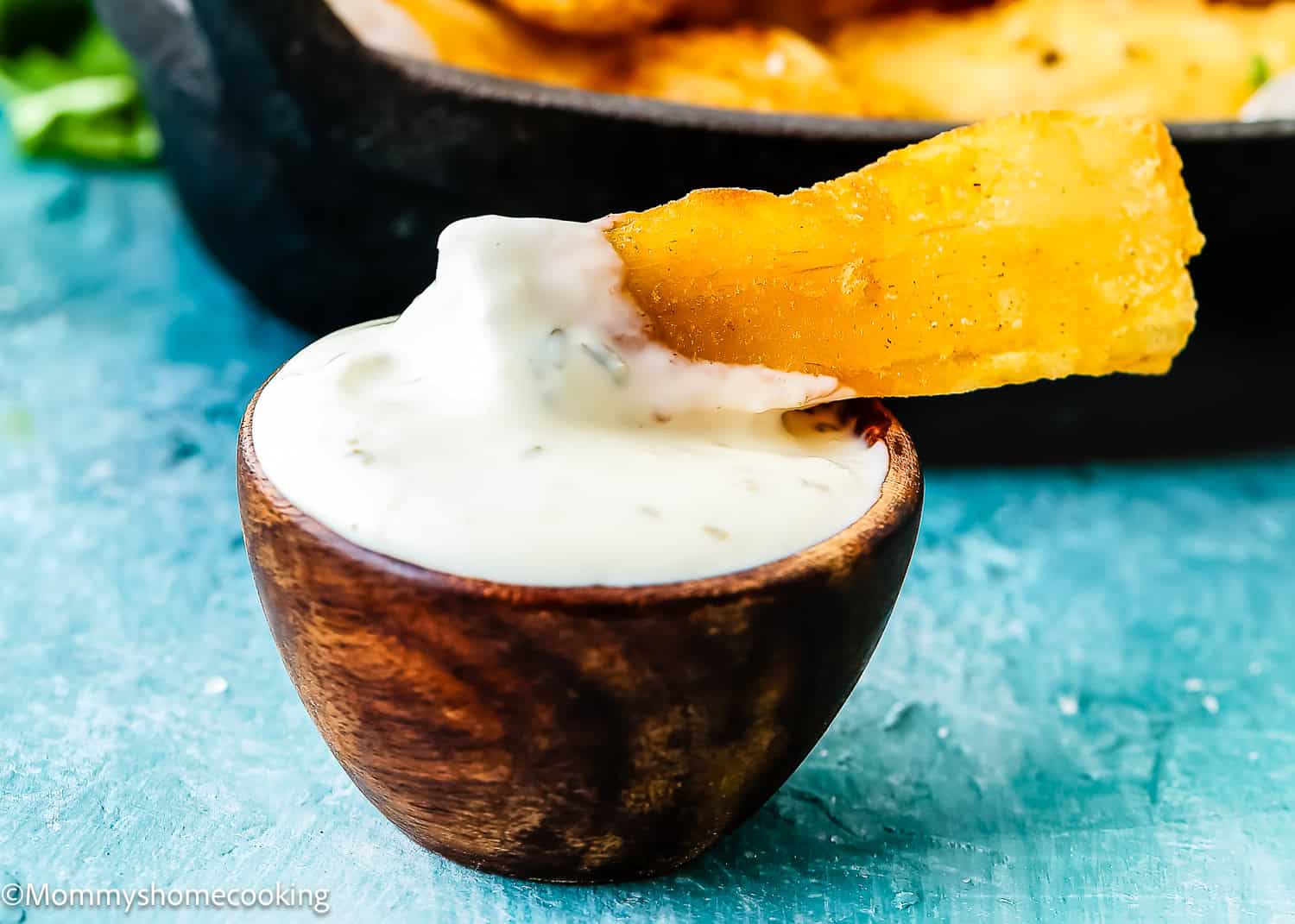 A fried yuca stick dipped in a small wooden bowl of creamy white sauce with a blurred background—discover how to make yuca fries for the perfect crispy snack.