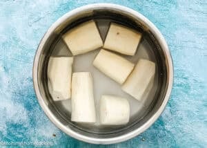 Chunks of peeled yuca root soaking in water inside a stainless steel bowl on a blue textured surface, a key step in how to make yuca fries.