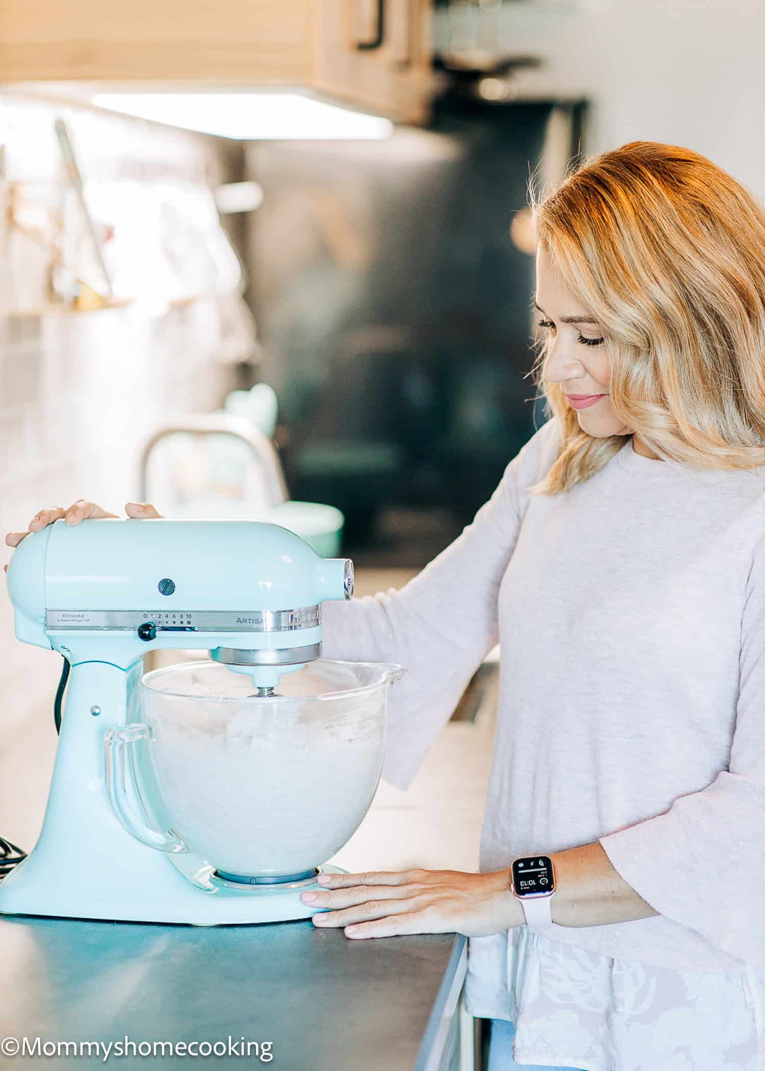 Oriana stands in a kitchen using a light blue stand mixer to blend ingredients in a glass bowl, mixing an egg-free cake.