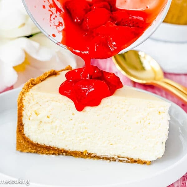 A slice of the Best Eggless Cheesecake on a plate is being topped with bright red strawberry sauce from a bowl.
