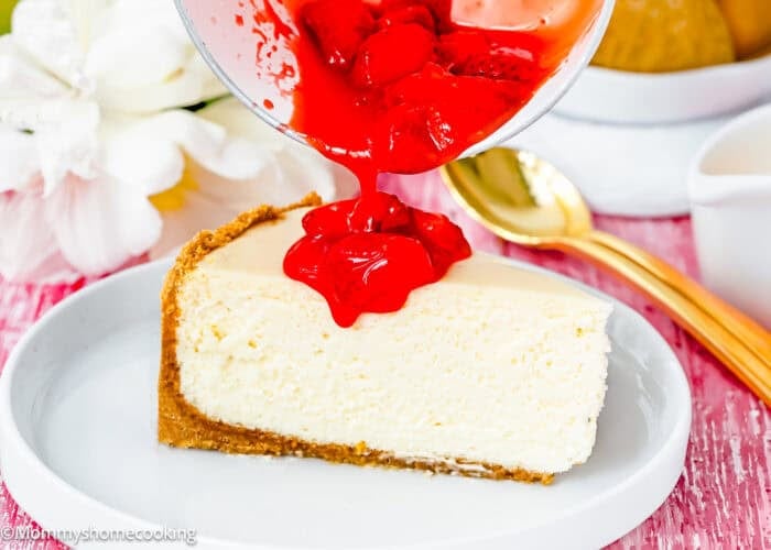 A slice of the Best Eggless Cheesecake on a plate is being topped with bright red strawberry sauce from a bowl.