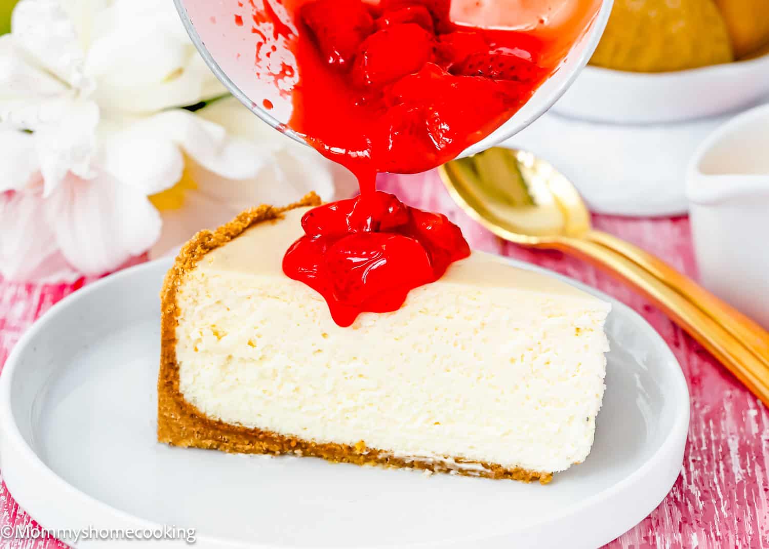 A slice of the Best Eggless Cheesecake on a plate is being topped with bright red strawberry sauce from a bowl.