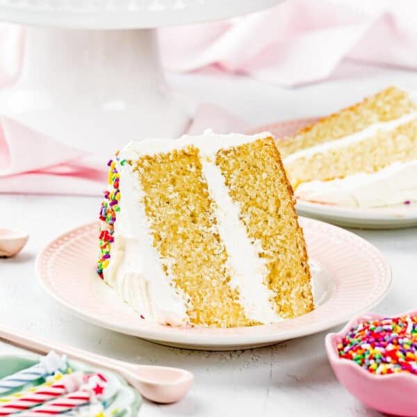 A slice of Egg-Free Vanilla Cake with white frosting and sprinkles is served on a pink plate; the rest of this soft, fluffy birthday cake without eggs sits on a stand in the background.