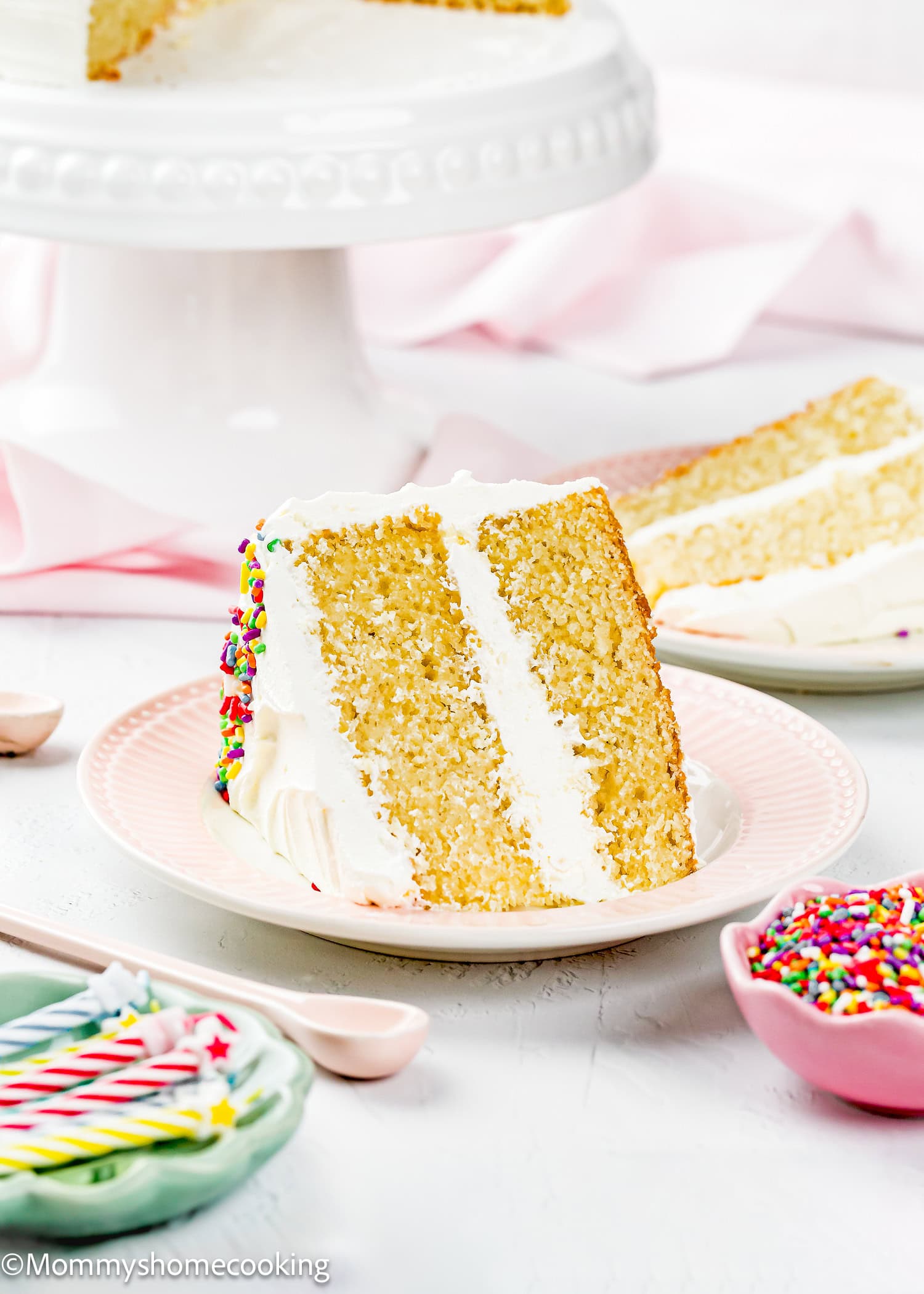 A slice of Egg-Free Vanilla Cake with white frosting and sprinkles is served on a pink plate; the rest of this soft, fluffy birthday cake without eggs sits on a stand in the background.