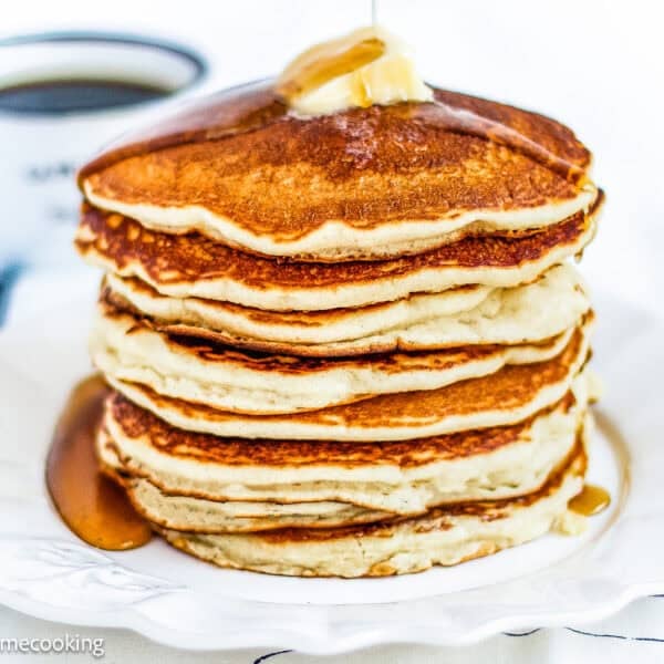 A stack of golden, fluffy eggless pancakes topped with a pat of butter and syrup on a white plate, with a cup of coffee in the background.