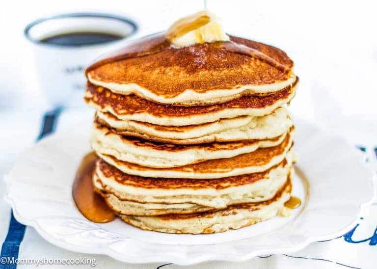 A stack of golden, fluffy eggless pancakes topped with a pat of butter and syrup on a white plate, with a cup of coffee in the background.
