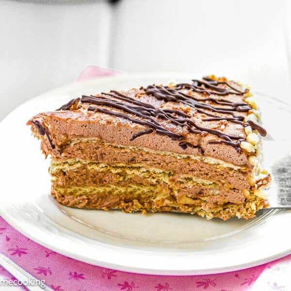 A slice of Marquesa de Chocolate, a Venezuelan no-bake chocolate cake with cream filling and chocolate drizzle, sits on a white plate with a cake server and spoon nearby.