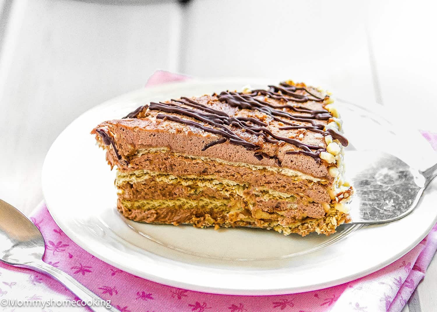 A slice of Marquesa de Chocolate, a Venezuelan no-bake chocolate cake with cream filling and chocolate drizzle, sits on a white plate with a cake server and spoon nearby.
