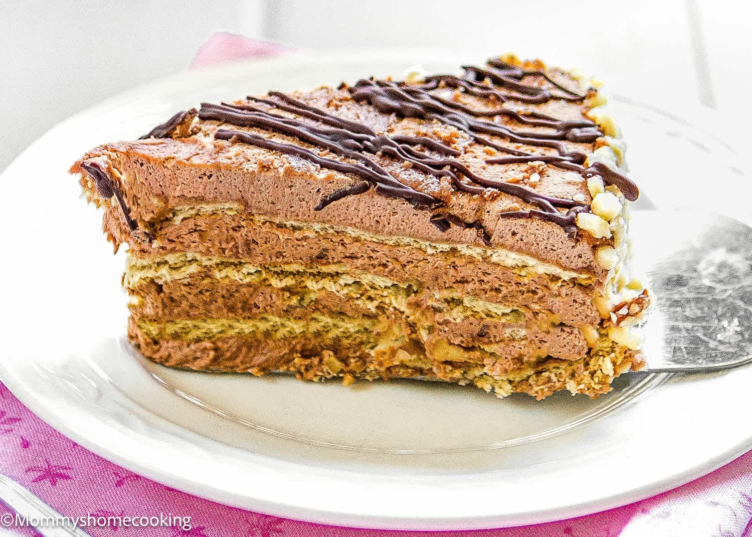 A slice of layered Marquesa de Chocolate (Venezuelan No-Bake Chocolate Cake) with cream filling and chocolate drizzle rests on a white plate, a cake server partially tucked beneath the decadent dessert.