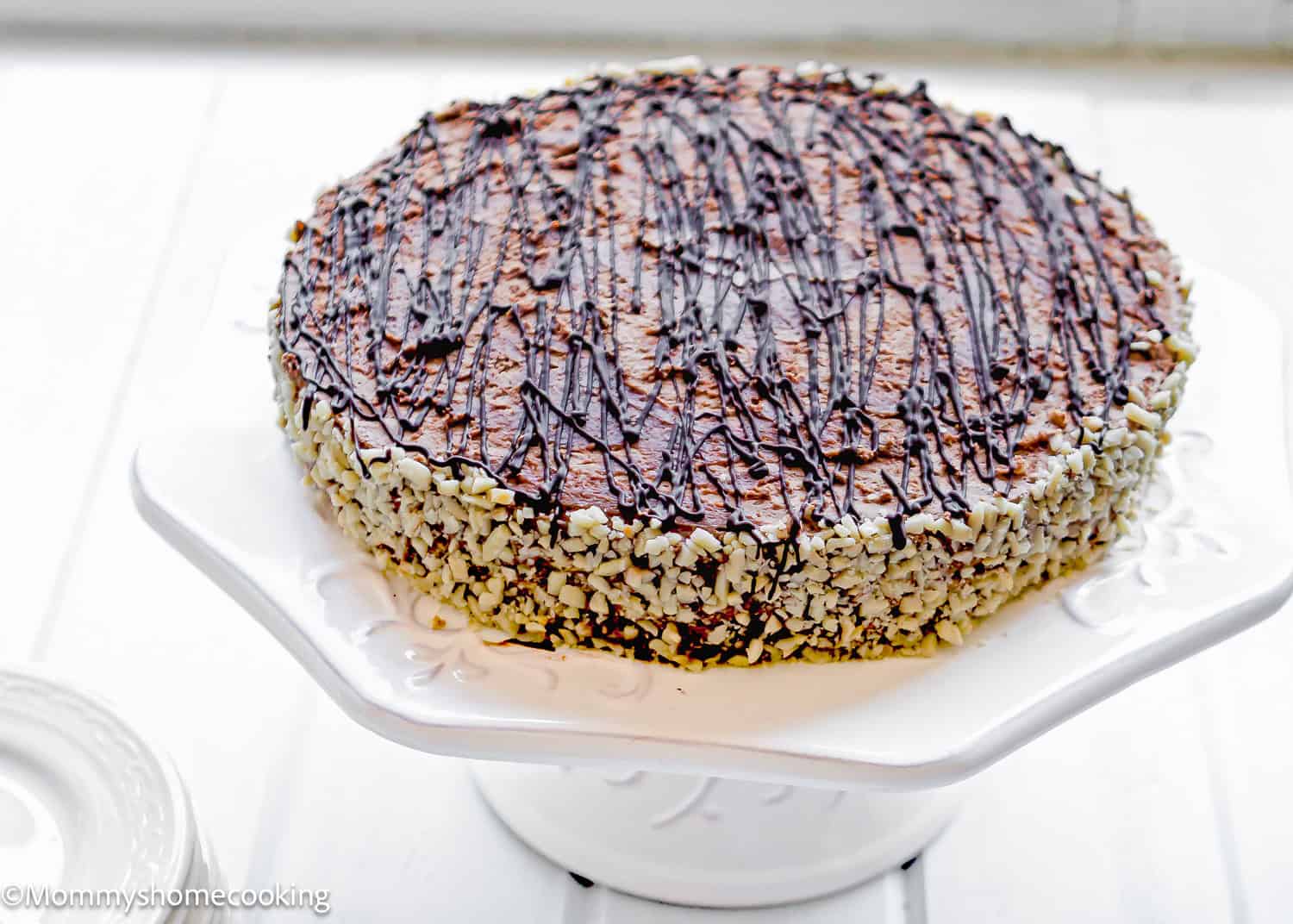 A round Marquesa de Chocolate (Venezuelan No-Bake Chocolate Cake) sits on a white stand, topped with a drizzle of chocolate and chopped nuts covering the sides.