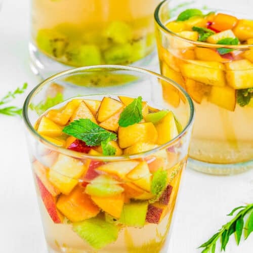 Three glasses filled with Easy Fruity White Sangria and assorted chopped fruits, including peaches, kiwi, and mint leaves, on a white surface.