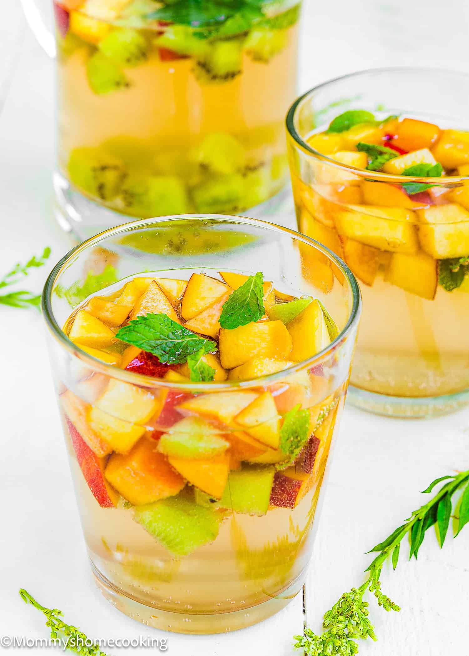 Three clear glasses filled with Easy Fruity White Sangria, diced fresh fruit, and mint leaves are set on a white surface with green leaves as decoration.