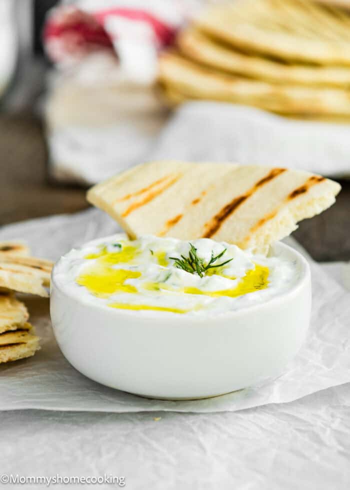 A bowl of easy Tzatziki sauce, featuring yogurt, drizzled with olive oil and sprinkled with dill, served alongside grilled flatbread pieces.