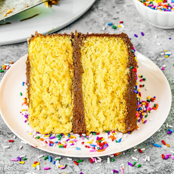 A slice of yellow layer cake made with a box mix without eggs, topped with chocolate frosting and colorful sprinkles, sits on a white plate with a fork and a bowl of extra sprinkles nearby.