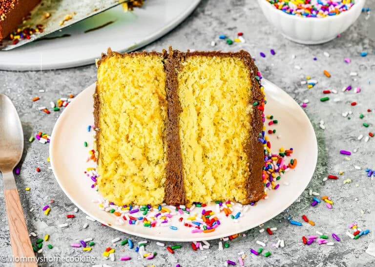 A slice of yellow layer cake made with a box mix without eggs, topped with chocolate frosting and colorful sprinkles, sits on a white plate with a fork and a bowl of extra sprinkles nearby.